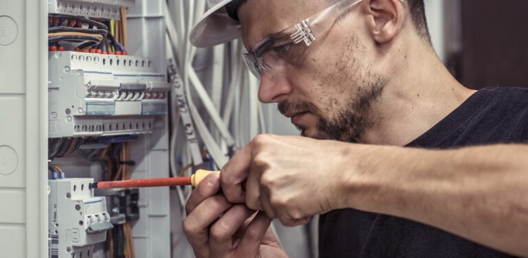 A male electrician works on a switchboard with electrical connecting cables, connecting equipment with tools in a complex job.